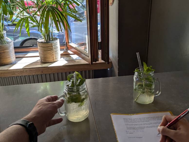 Picture taken in a restaurant. One hand is signing a written contract. There are two lemonade glasses on the table. In the background, green plants mostly hide the view outside the window.